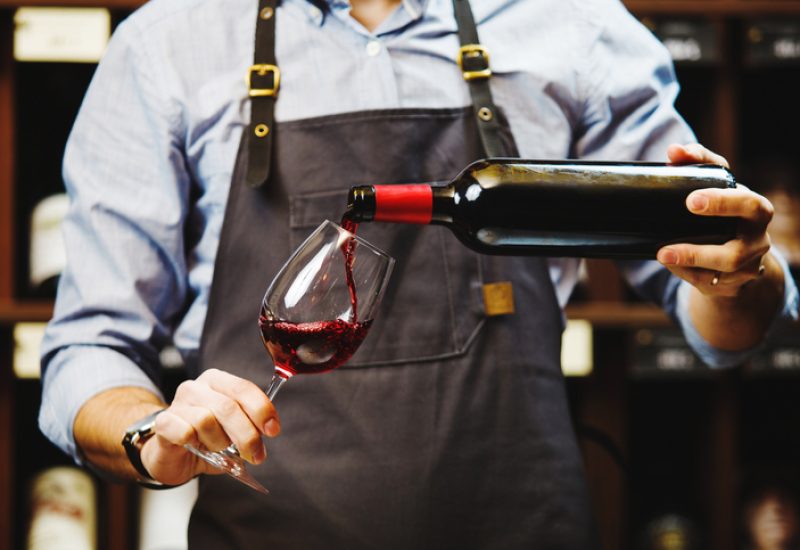 Male sommelier pouring red wine into long-stemmed wineglasses. Waiter with bottle of alcohol beverage. Bartender at bar counter pour elite drink into long-stemmed glass