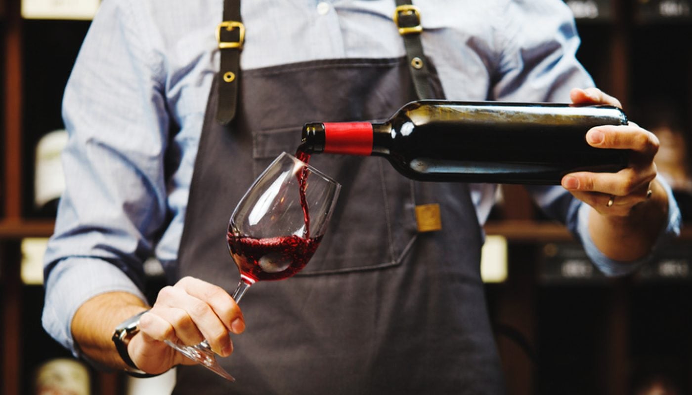 Male sommelier pouring red wine into long-stemmed wineglasses. Waiter with bottle of alcohol beverage. Bartender at bar counter pour elite drink into long-stemmed glass